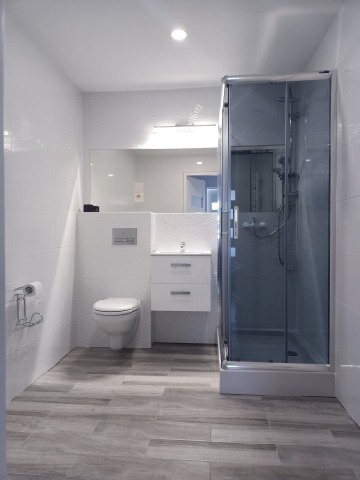 Jasna łazienka z kabiną prysznicową, toaletą podwieszaną, umywalką z szafką i podłogą z płytek imitujących drewno, oświetlenie sufitowe i nad lustrem.