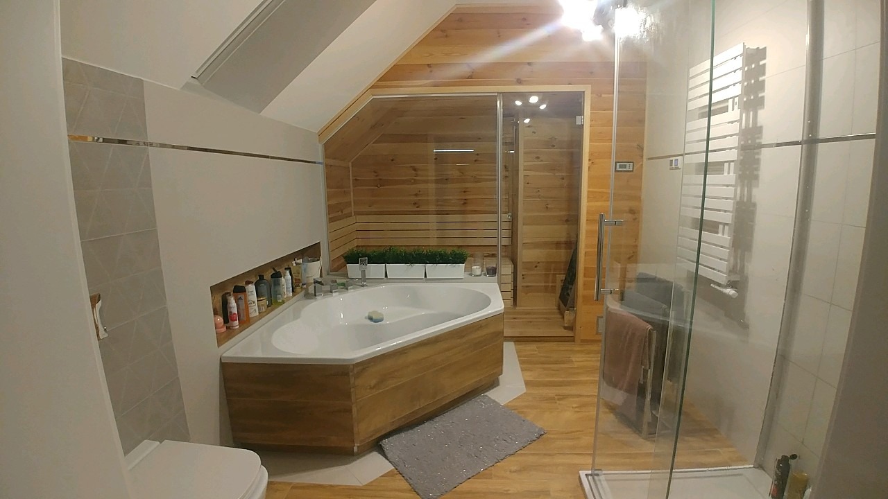 Nowoczesna łazienka na poddaszu z drewnianymi panelami, narożną wanną obudowaną drewnem i szklaną kabiną prysznicową.