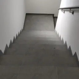 Schody wyłożone szarą, matową glazurą, widok z g&oacute;ry, z minimalistyczną metalową poręczą po prawej stronie, białe ściany z charakterystycznym wykończeniem krawędzi.