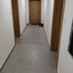 SANAN GROUP - Korytarz z szarymi płytkami podłogowymi, trzema drewnianymi drzwiami i kablem elektrycznym leżącym na podłodze.