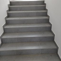 SANAN GROUP - Schody wyłożone szarymi, matowymi płytkami ceramicznymi, prowadzące do metalowej bramy z widokiem na budynek w tle.