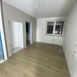 SANAN GROUP - Pokój w trakcie remontu: świeżo położona podłoga panelowa, ściany częściowo pomalowane na szaro, widoczne instalacje elektryczne i hydrauliczne, przygotowane otwory drzwiowe z niebieską taśmą...