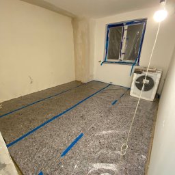 SANAN GROUP - Przygotowanie pokoju do prac wykończeniowych: podłoga zabezpieczona folią i taśmą malarską, okno zakryte folią ochronną, widoczny klimatyzator przenośny.