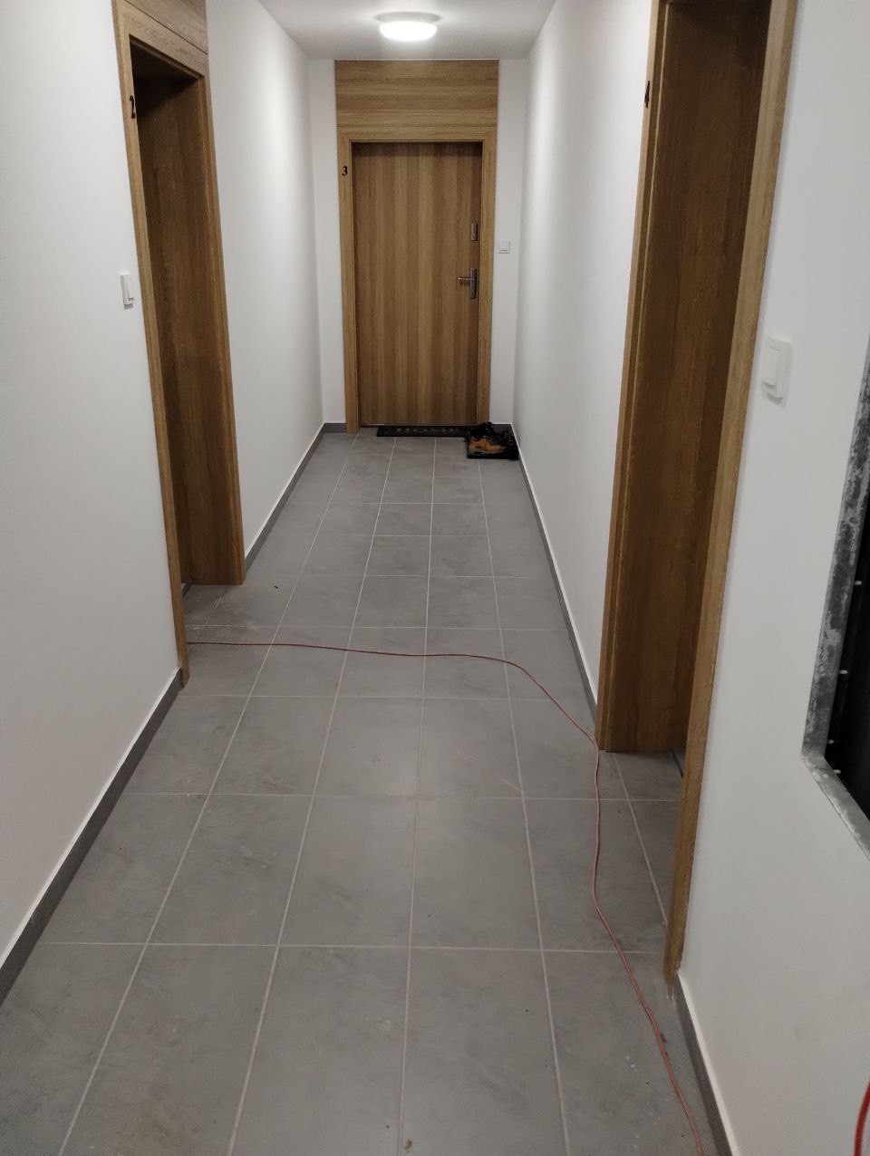 Korytarz z szarymi płytkami podłogowymi, trzema drewnianymi drzwiami i kablem elektrycznym leżącym na podłodze.