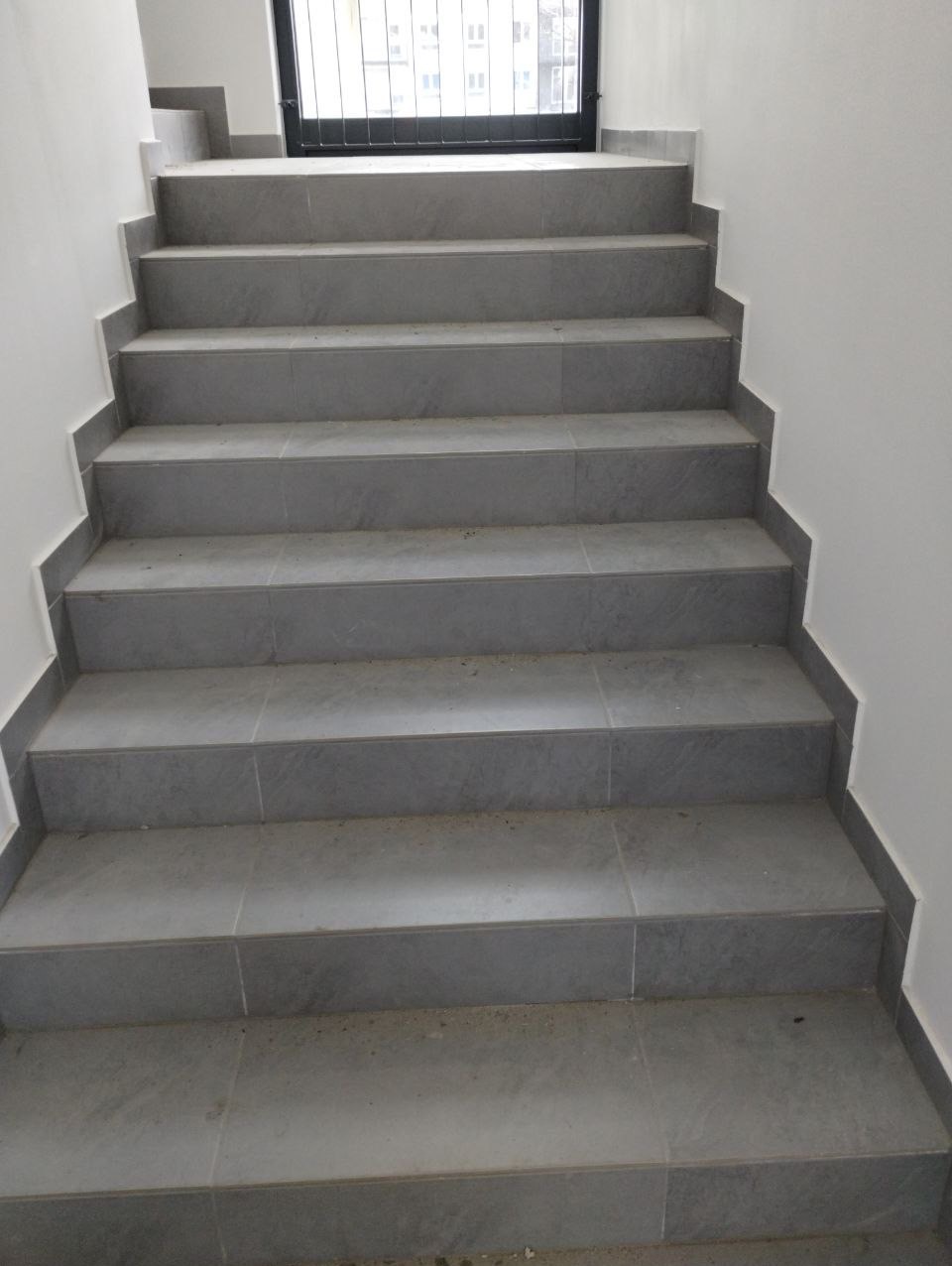 Schody wyłożone szarymi, matowymi płytkami ceramicznymi, prowadzące do metalowej bramy z widokiem na budynek w tle.