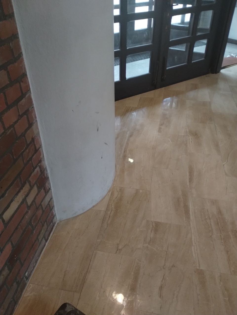 Beżowa, marmurkowa podłoga z połyskiem, przylegająca do ceglanej ściany i zaokrąglonego, białego filaru, w tle ciemne, przeszklone drzwi.