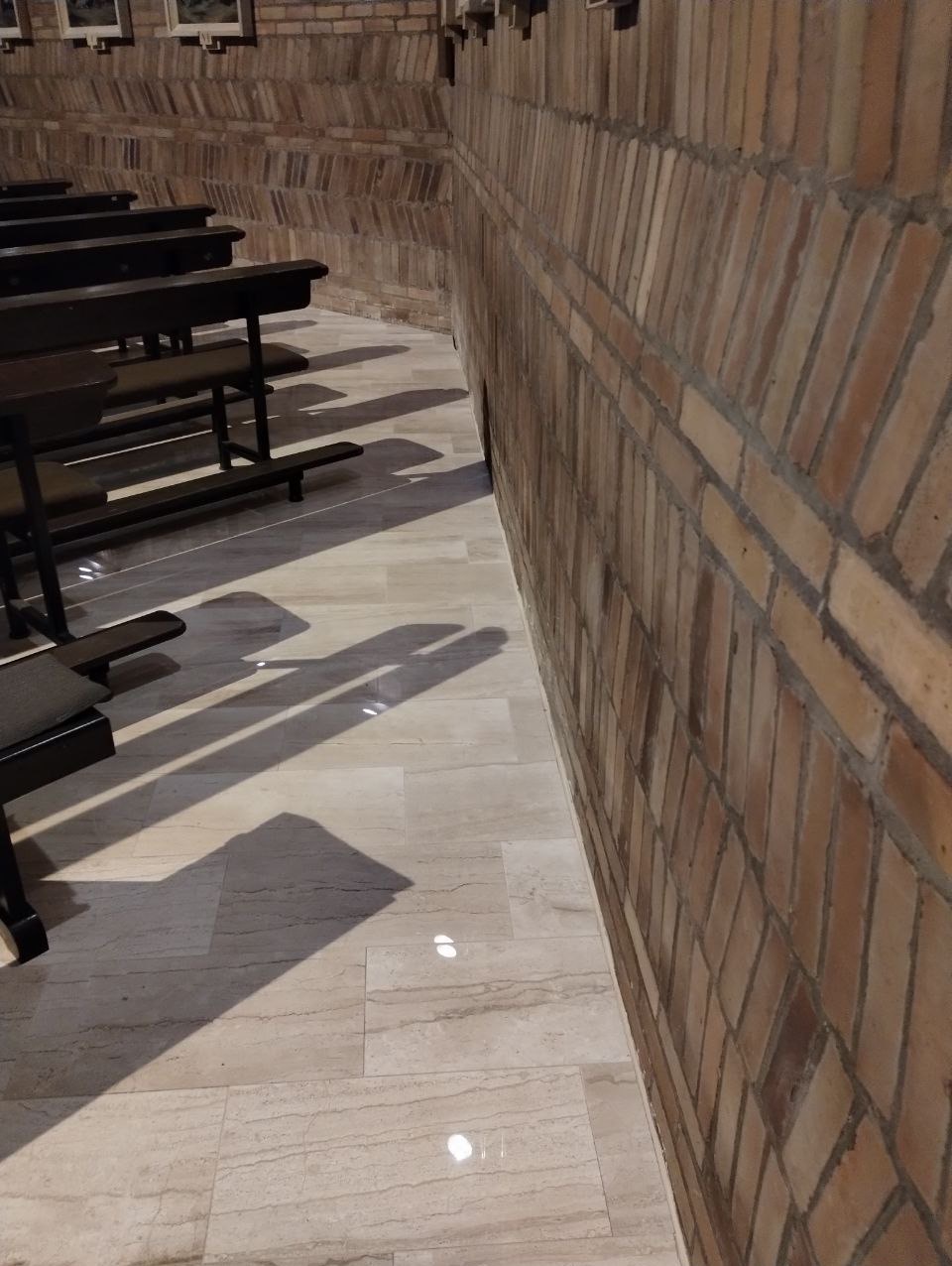 Zakrzywiona ściana z cegły w odcieniach brązu sąsiaduje z podłogą wyłożoną jasnymi płytkami imitującymi marmur, rzucając cień ławek wzdłuż ściany.