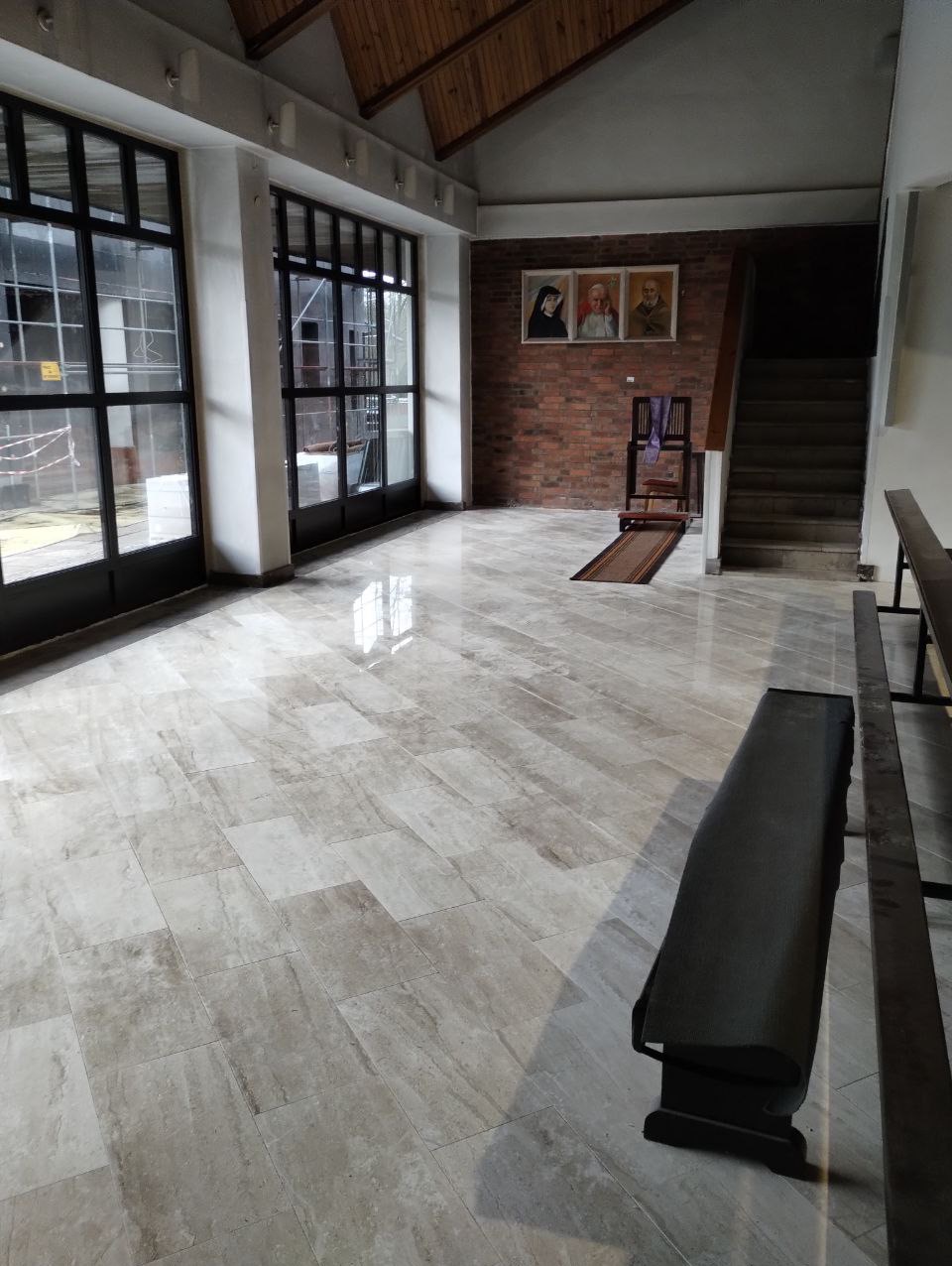 Jasne, polerowane płytki podłogowe ułożone w dużej sali z wysokimi oknami i ceglaną ścianą z portretami, widoczna ławka i schody.