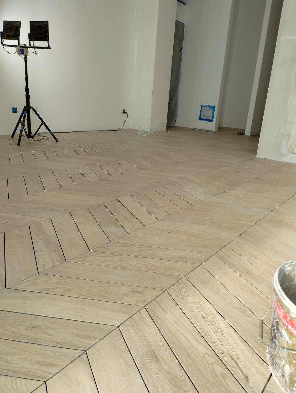 Jasne, drewnopodobne płytki ułożone we wzór jodełki w przestronnym pomieszczeniu z białymi ścianami, oświetlone lampami budowlanymi na statywie, widoczny fragment wiadra przy krawędzi kadru.