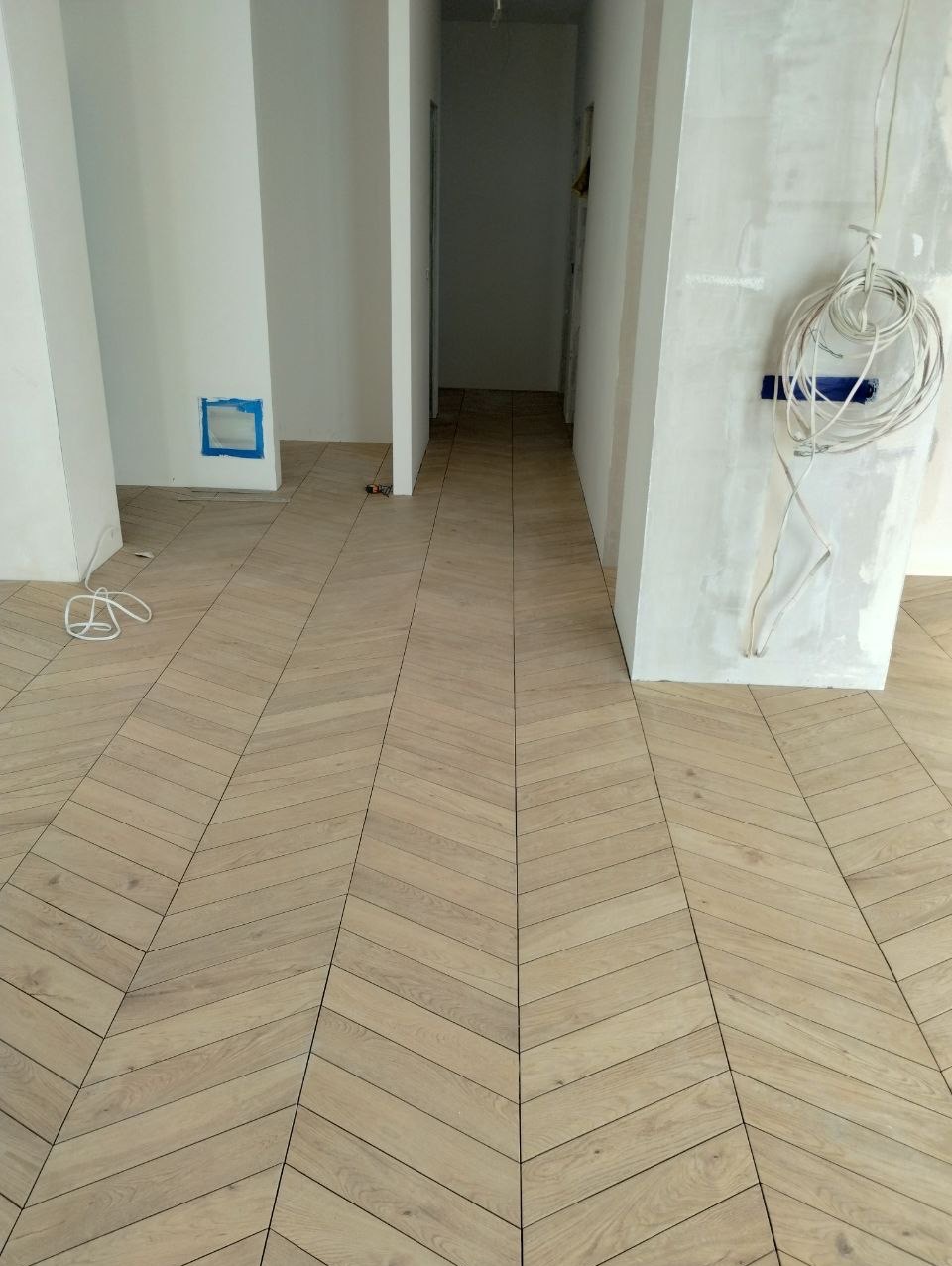 Ułożona jasna podłoga z płytek w jodełkę w korytarzu, widoczne kable i niedokończone ściany w trakcie remontu.