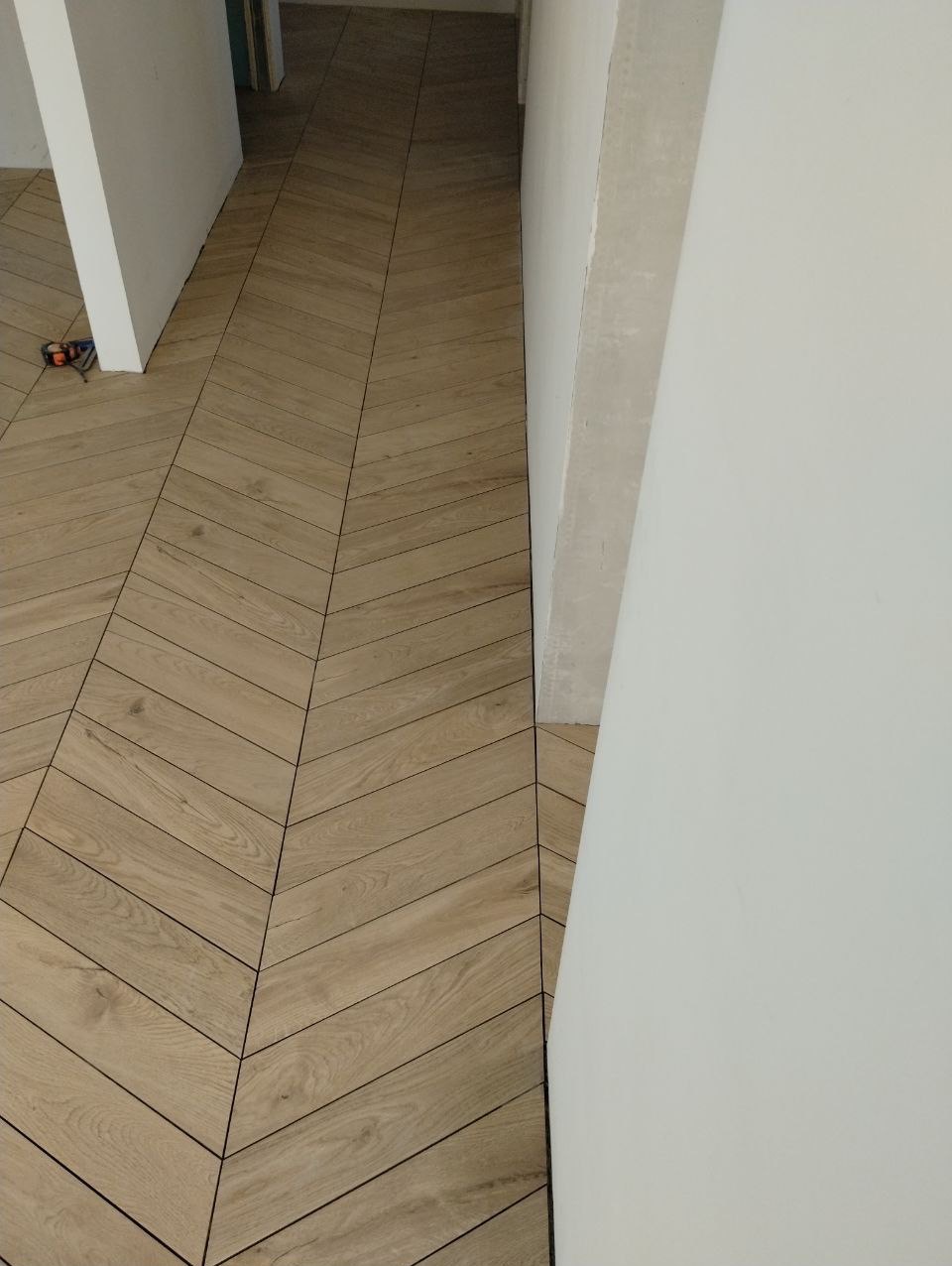 Ułożona w jodełkę podłoga z płytek imitujących drewno, widok z góry, fragment białej ściany po prawej stronie kadru.