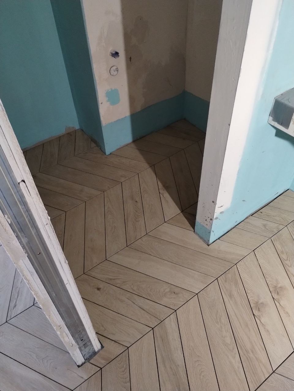 Ułożona pod kątem jodełki jasna terakota imitująca drewno na podłodze w rogu pomieszczenia z niebieskimi ścianami, widoczny fragment futryny drzwi.