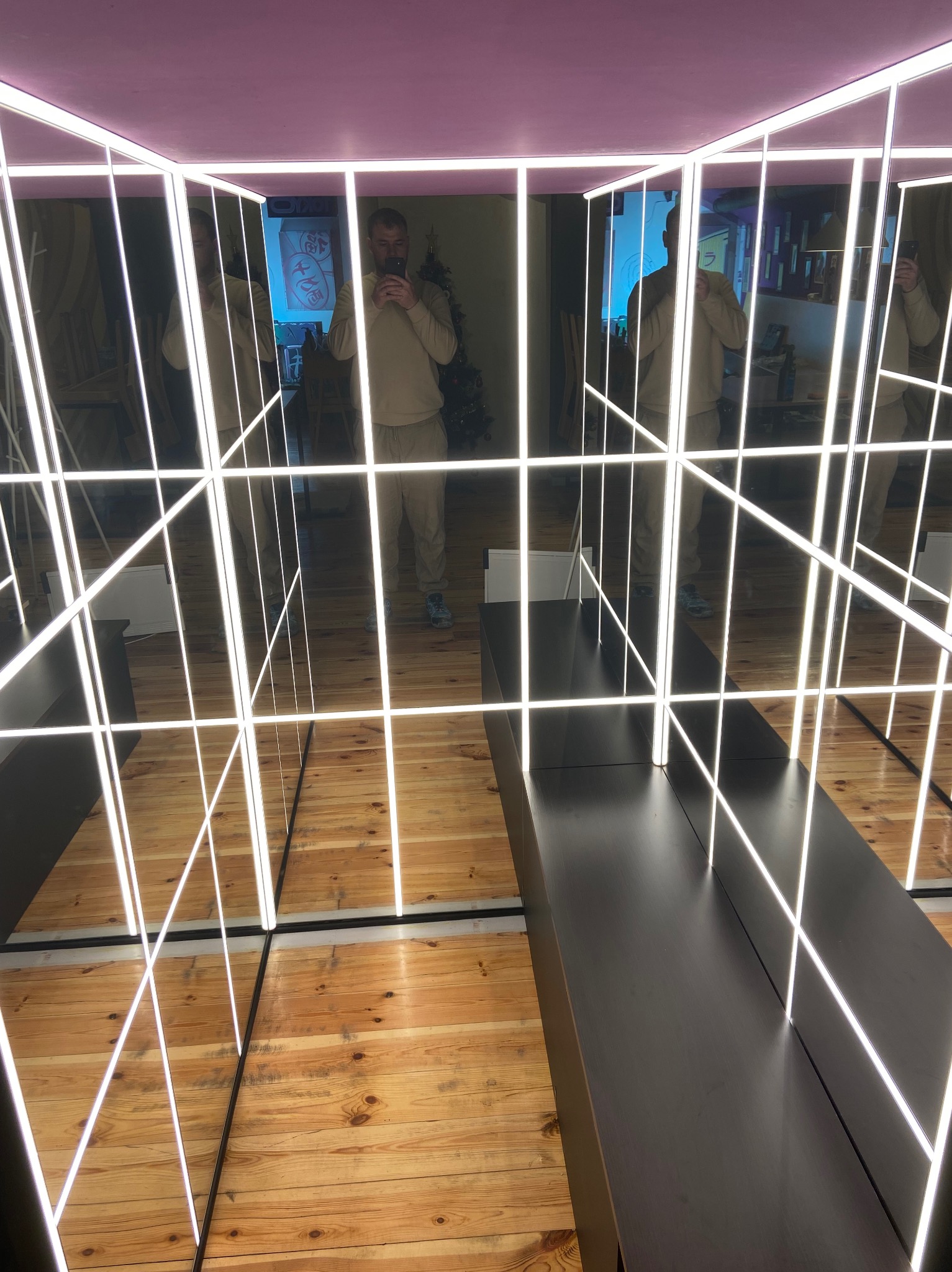 Instalacja artystyczna z lustrami i neonowymi światłami na drewnianej podłodze, odbicie osoby robiącej zdjęcie telefonem.