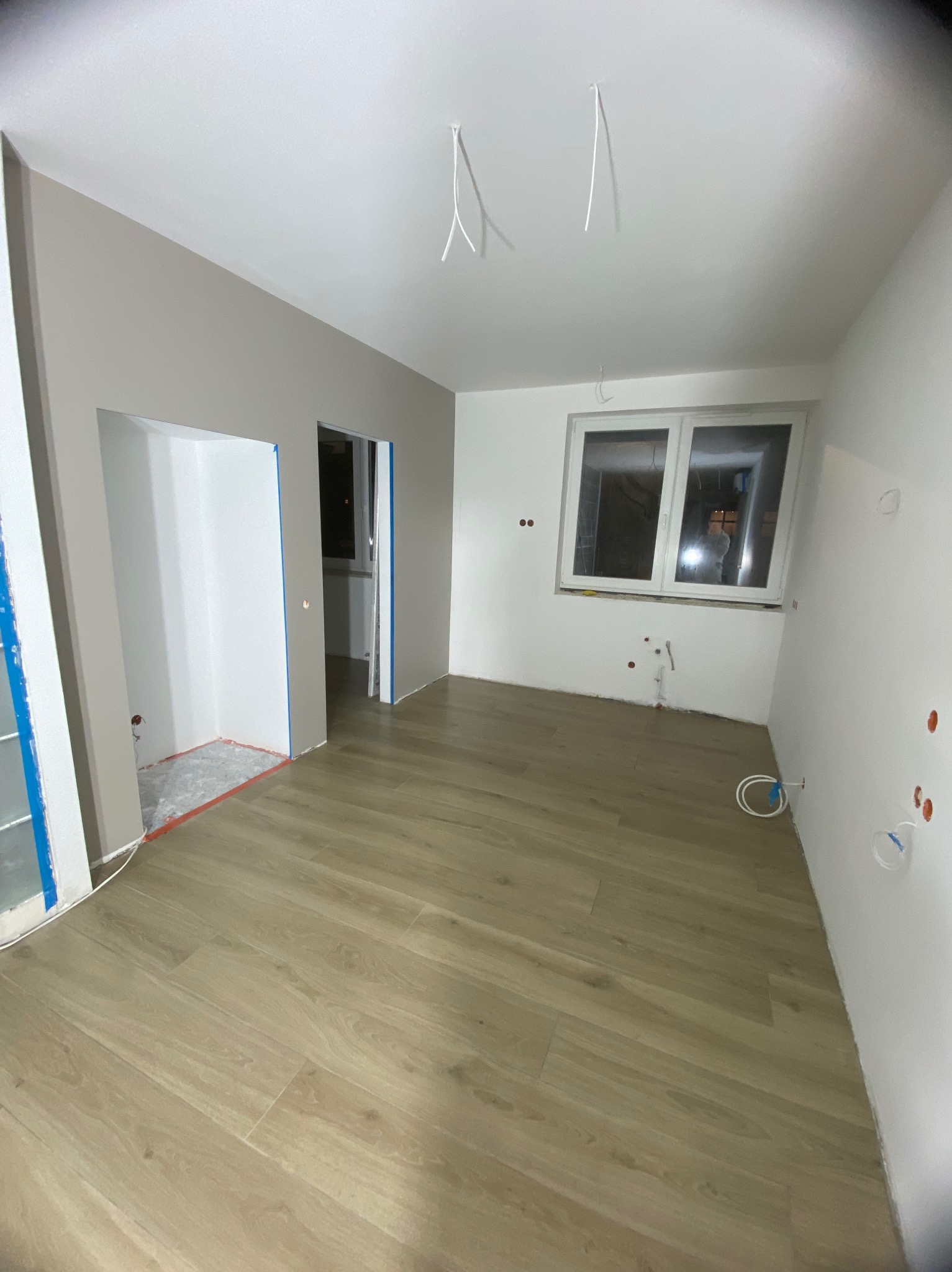 Pokój w trakcie remontu: świeżo położona podłoga panelowa, ściany częściowo pomalowane na szaro, widoczne instalacje elektryczne i hydrauliczne, przygotowane otwory drzwiowe z niebieską taśmą...