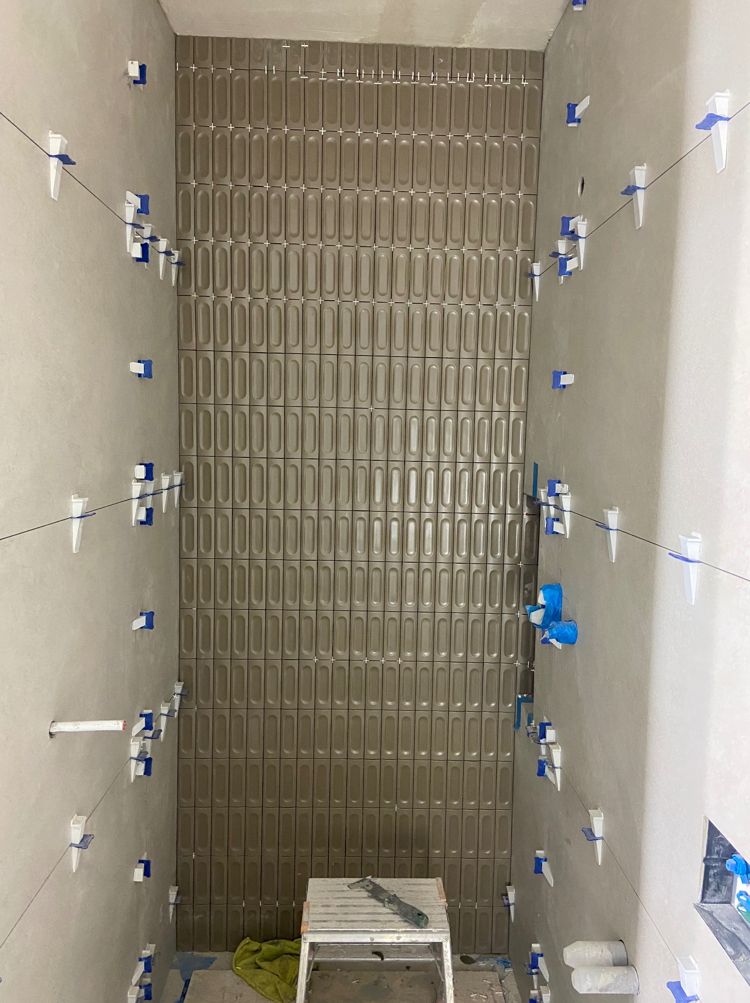 Montaż podłużnych, brązowych płytek w układzie pionowym z użyciem systemu poziomowania płytek w łazience w Katowicach. Widoczny fragment ściany z zamontowanymi płytkami oraz narzędzia na małym...