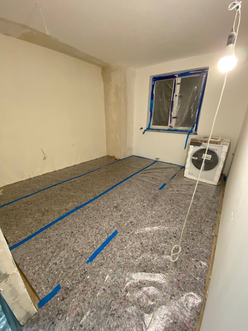 Przygotowanie pokoju do prac wykończeniowych: podłoga zabezpieczona folią i taśmą malarską, okno zakryte folią ochronną, widoczny klimatyzator przenośny.