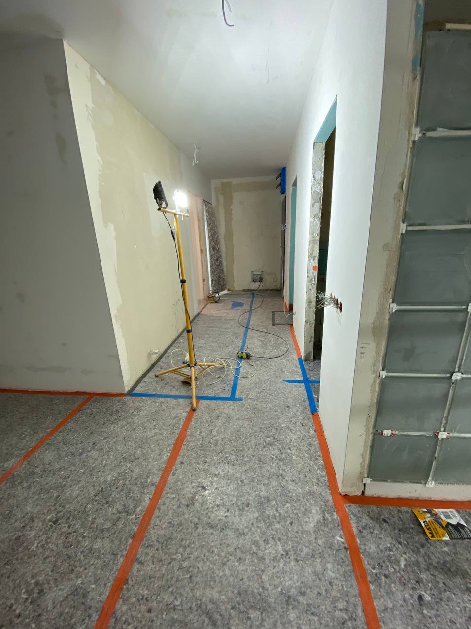 Korytarz w trakcie prac przygotowawczych: podłoga zabezpieczona szarą matą z pomarańczowymi liniami, ściany częściowo wyszpachlowane, widoczna lampa na statywie i niebieska taśma malarska.