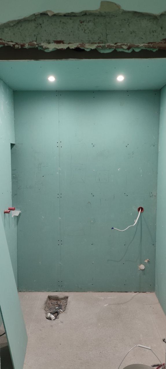 Wnętrze łazienki w trakcie remontu: ściany wyłożone płytami gipsowo-kartonowymi, widoczne instalacje elektryczne i hydrauliczne, otwór w podłodze.