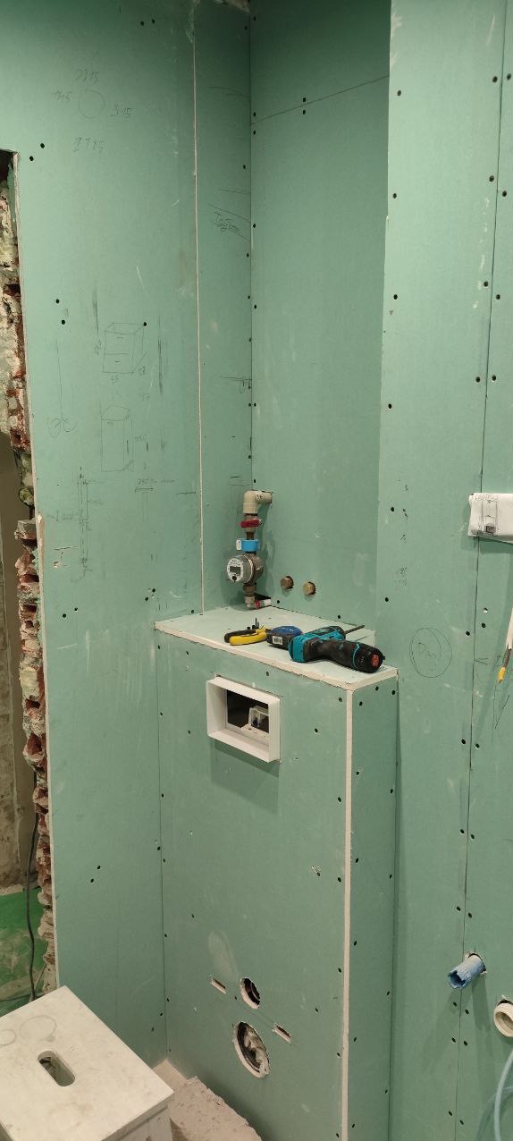 Ściany w łazience pokryte zielonymi płytami gipsowo-kartonowymi, widoczne ślady ołówka z naniesionymi wymiarami, instalacja wodna z licznikiem, otwór na wentylację i narzędzia na półce.
