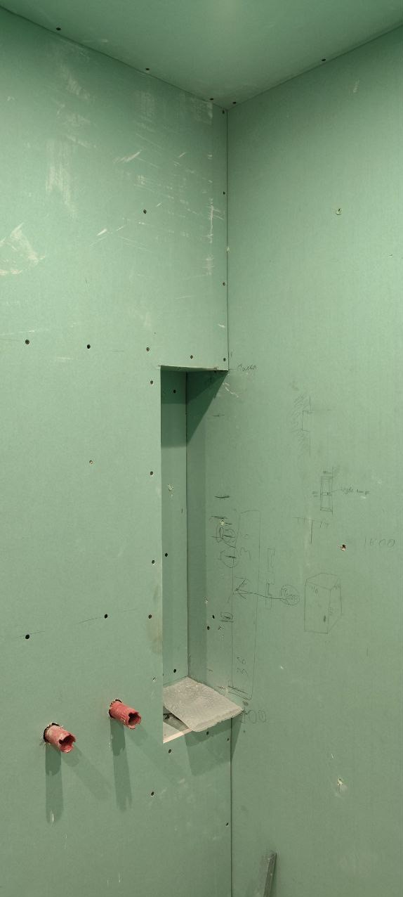 Ściana z płyt kartonowo-gipsowych w narożniku łazienki, z wnęką na półkę, widoczne wystające rury instalacji wodnej i odręczne szkice na ścianie.