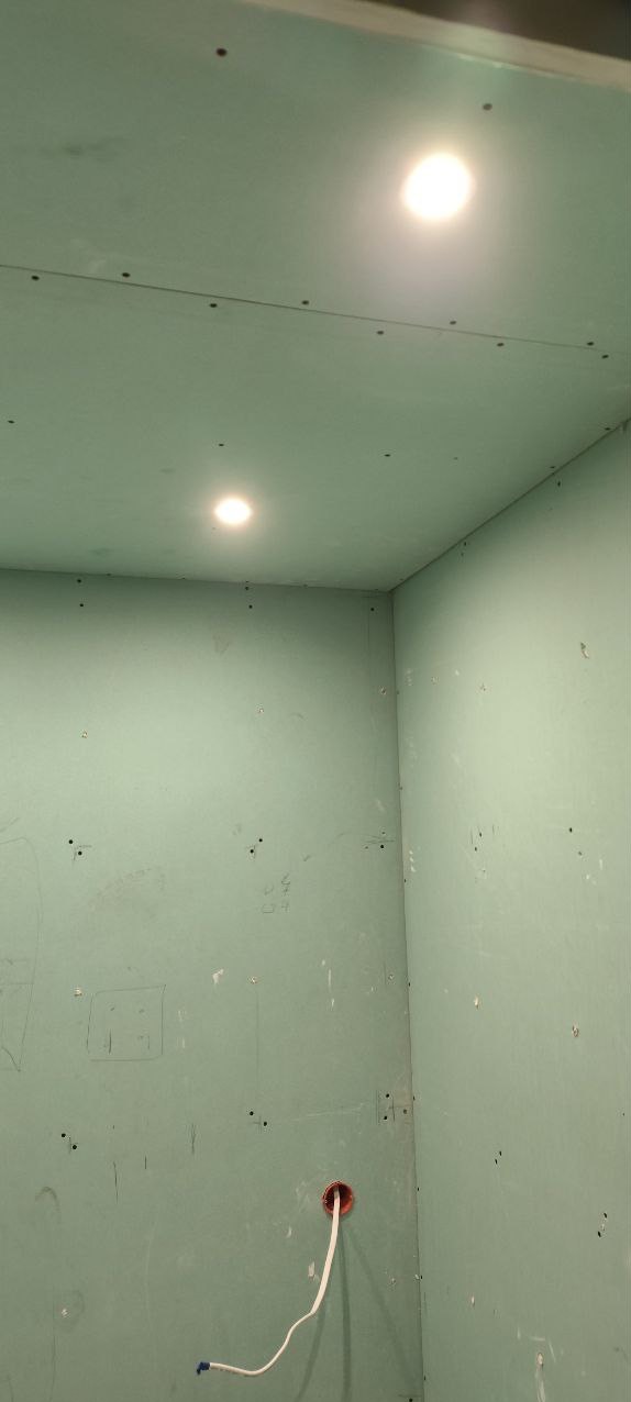 Wnętrze pokoju z zamontowanymi płytami gipsowo-kartonowymi, widoczne ślady ołówka na ścianach, otwór na gniazdko elektryczne z wystającym przewodem oraz dwa punkty oświetleniowe w suficie.