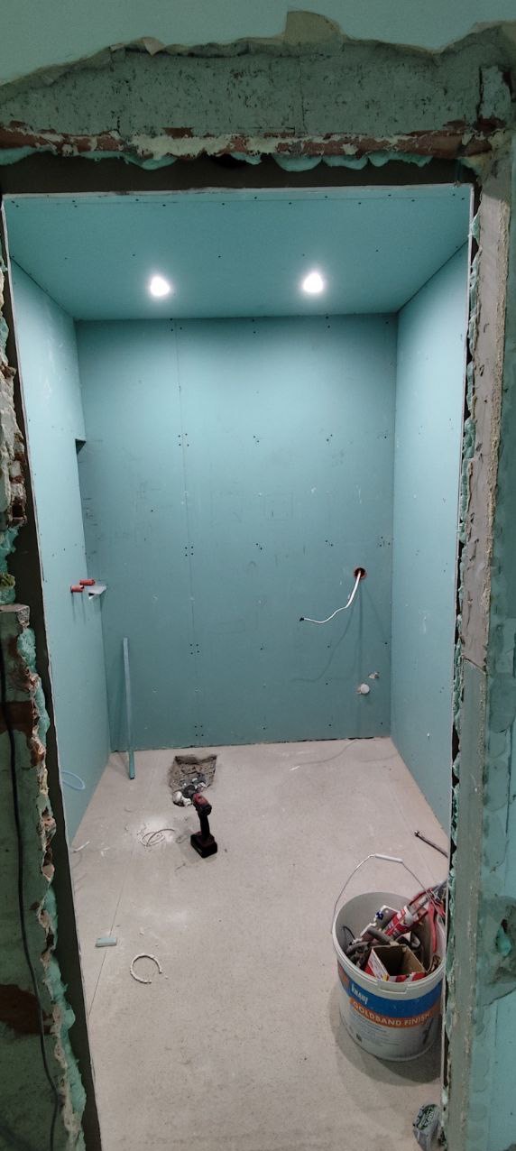 Łazienka w trakcie remontu: ściany i sufit pokryte niebieskimi płytami gipsowo-kartonowymi, widoczne otwory na oświetlenie, niezabezpieczone przewody, narzędzia i wiadro z materiałami na podłodze...