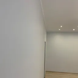 Świeżo wykończone wnętrze z pomalowanymi na szaro ścianami, jasnym parkietem i punktowym oświetleniem sufitowym; widoczny fragment futryny drzwi.