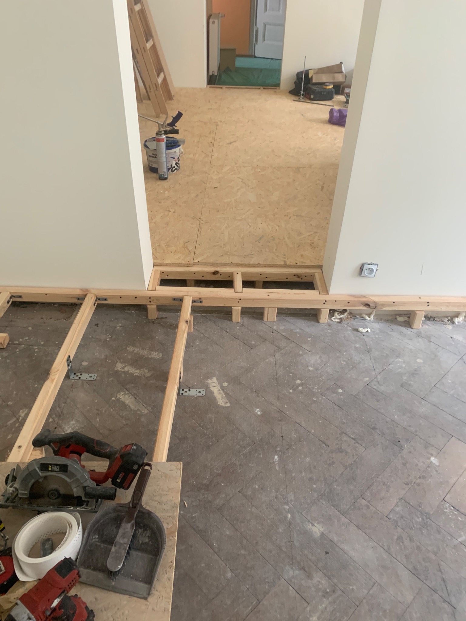 Przygotowanie podłoża pod nową podłogę: drewniana konstrukcja legarów na starej, ułożonej w jodełkę podłodze, narzędzia budowlane w pobliżu, widok na sąsiednie pomieszczenie z częściowo ułożoną...