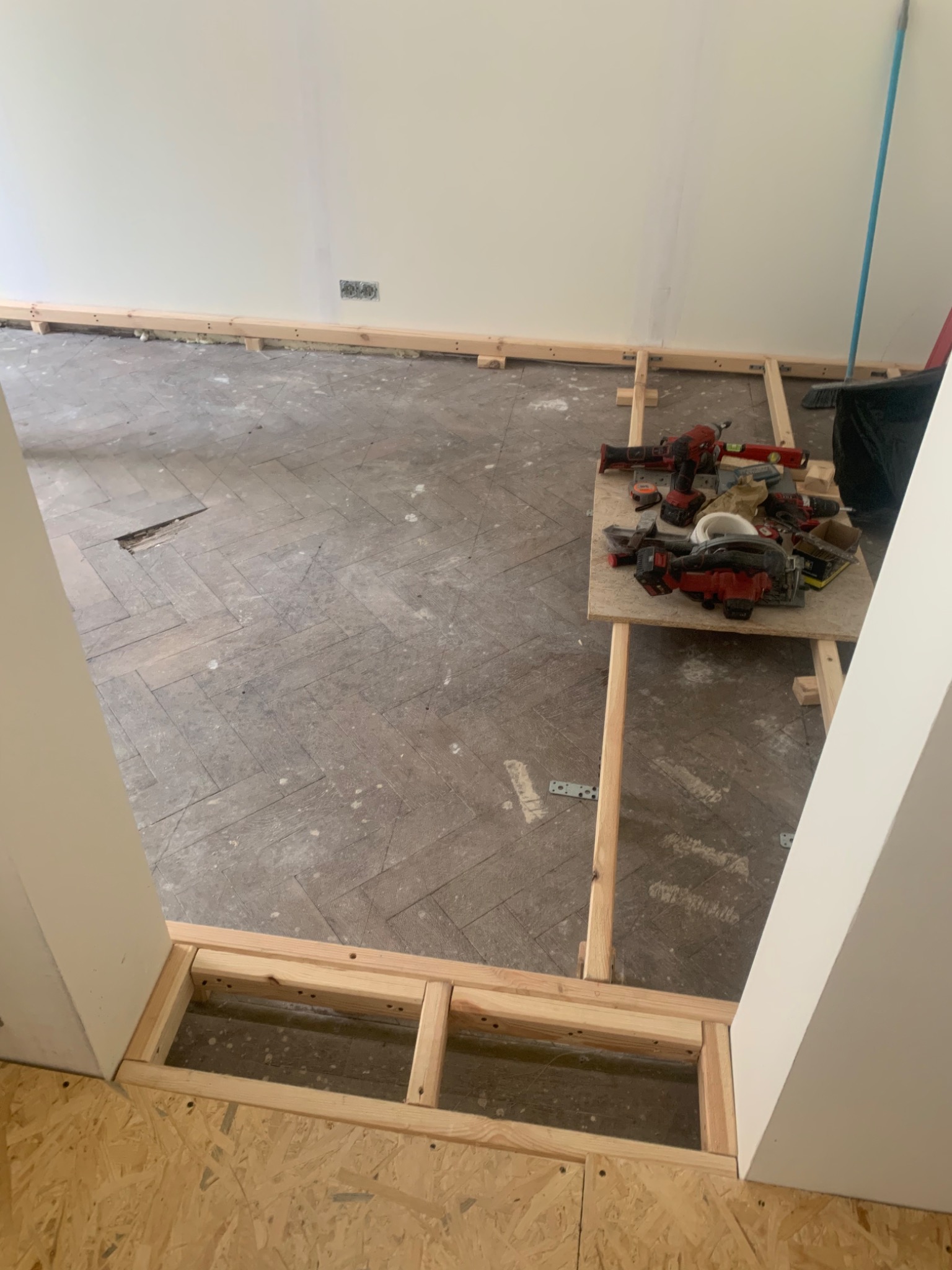 Przygotowanie do wymiany podłogi: widoczny fragment starego parkietu w jodełkę, drewniana konstrukcja pod podłogę przy progu oraz narzędzia na prowizorycznym stole roboczym.