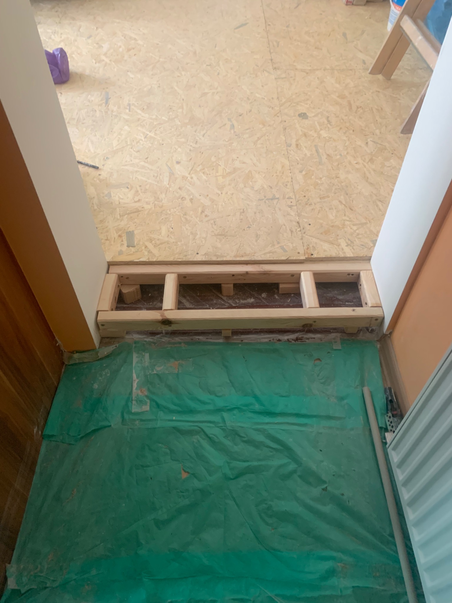 Przygotowanie podłoża pod podłogę: drewniana konstrukcja legarowa na zielonej folii ochronnej, widok z góry na surową płytę OSB i fragmenty ścian.