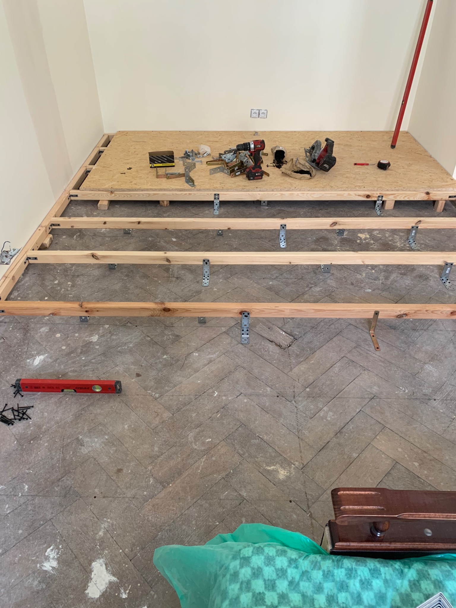 Konstrukcja podłogi z drewnianych legarów i płyty OSB, widoczne narzędzia, poziomica i walizka na podłodze w jodełkę, etap prac remontowych przed ułożeniem paneli.