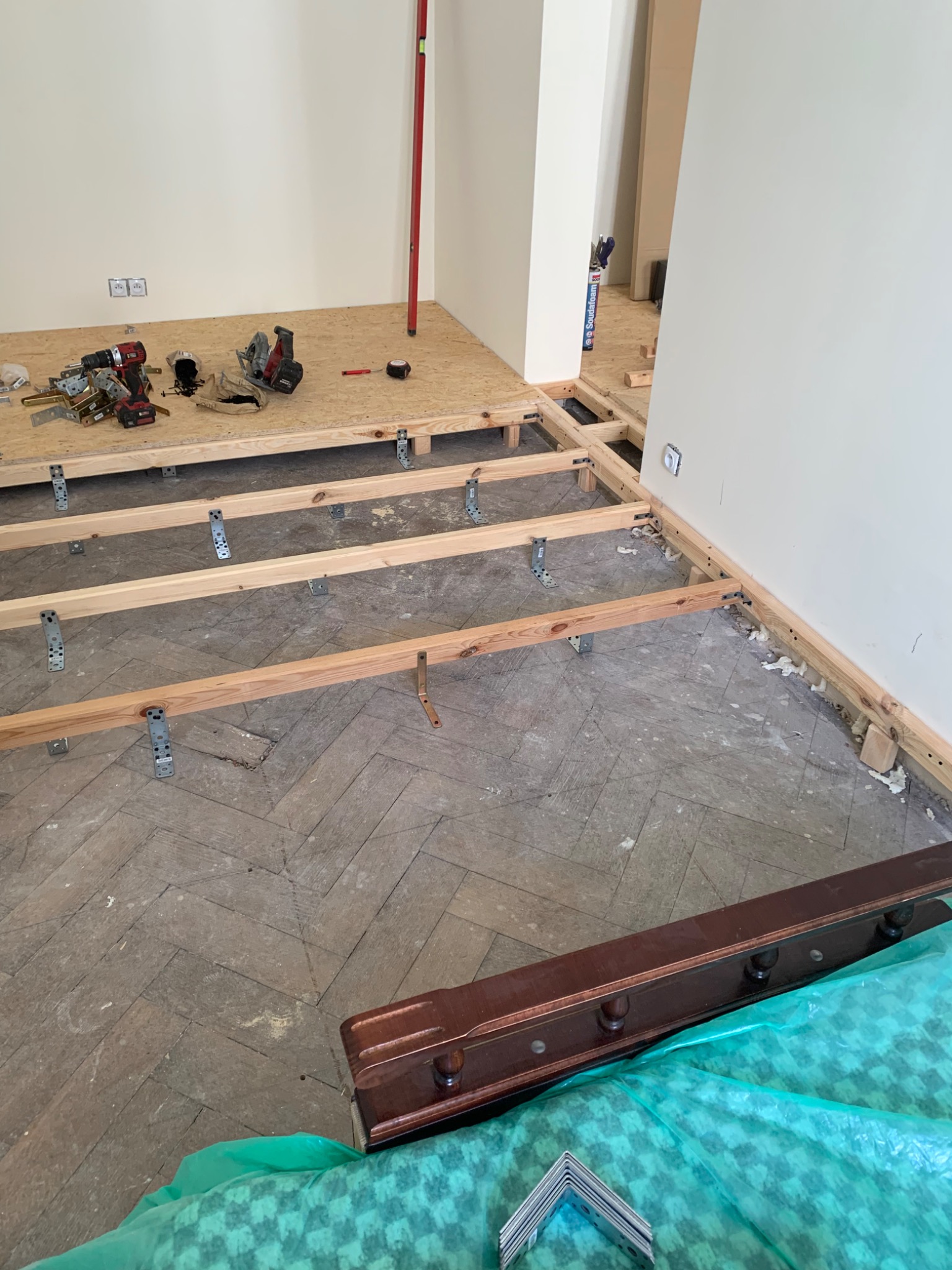 Przygotowanie podłoża pod nową podłogę: drewniana konstrukcja legarów na starym parkiecie ułożonym w jodełkę, widoczne narzędzia i poziomica, ściany przygotowane do dalszych prac remontowych.