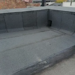 Płaski dach pokryty papą termozgrzewalną z widocznymi łączeniami i kominkiem odpowietrzającym w rogu, na tle zabudowy miejskiej.