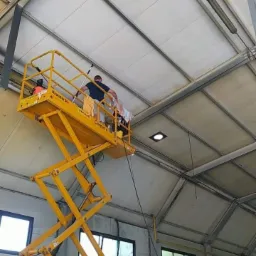 Pracownik na podnośniku koszowym czyści sufit hali przemysłowej, widoczne okna, drabina i elementy konstrukcyjne budynku.