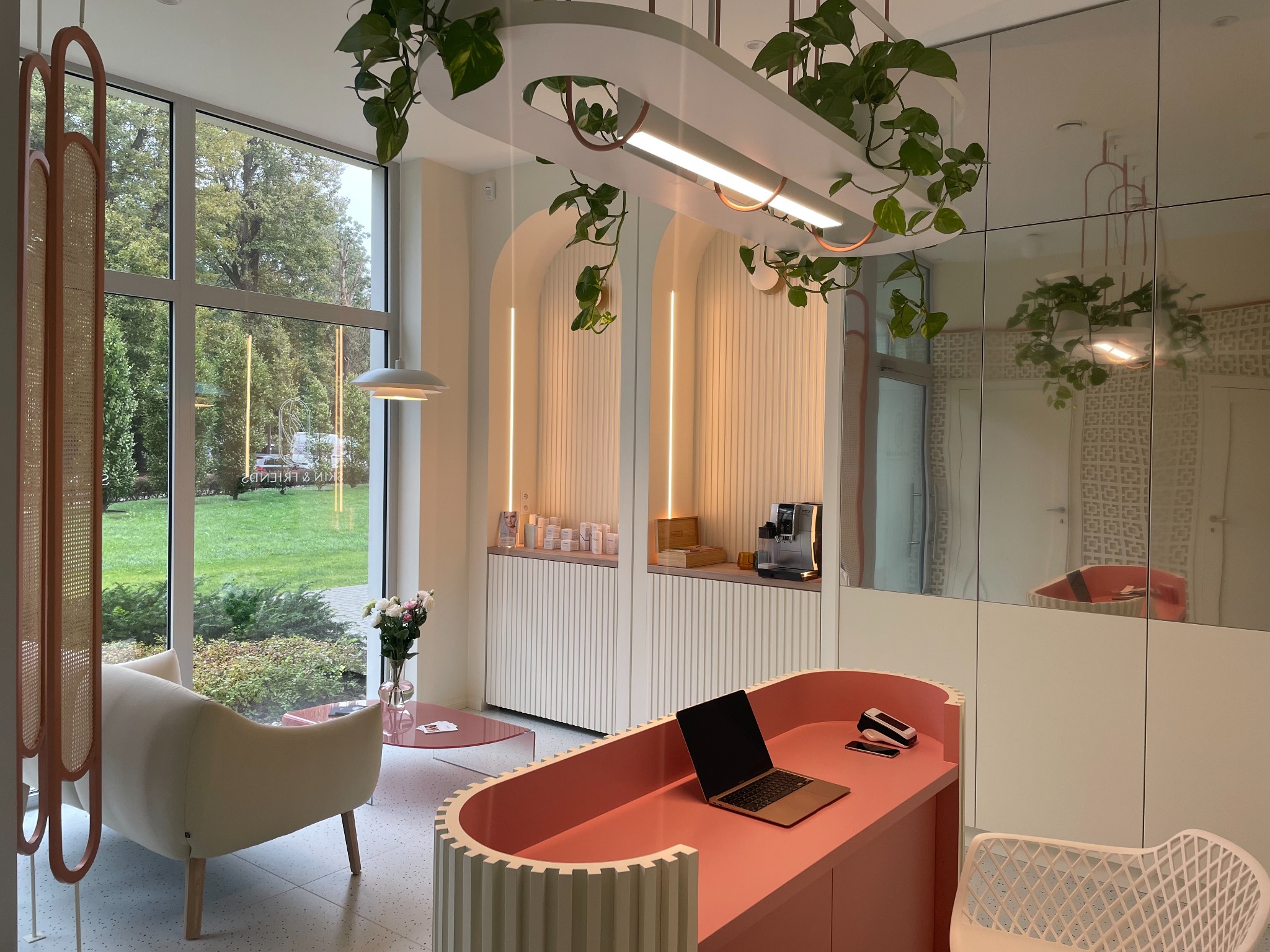 Jasne wnętrze recepcji z biurkiem w odcieniach koralowych, laptopem, terminalem płatniczym i telefonem, dekorowane roślinami zwisającymi z sufitu i kwiatami w wazonie, z widokiem na zieleń za oknem.