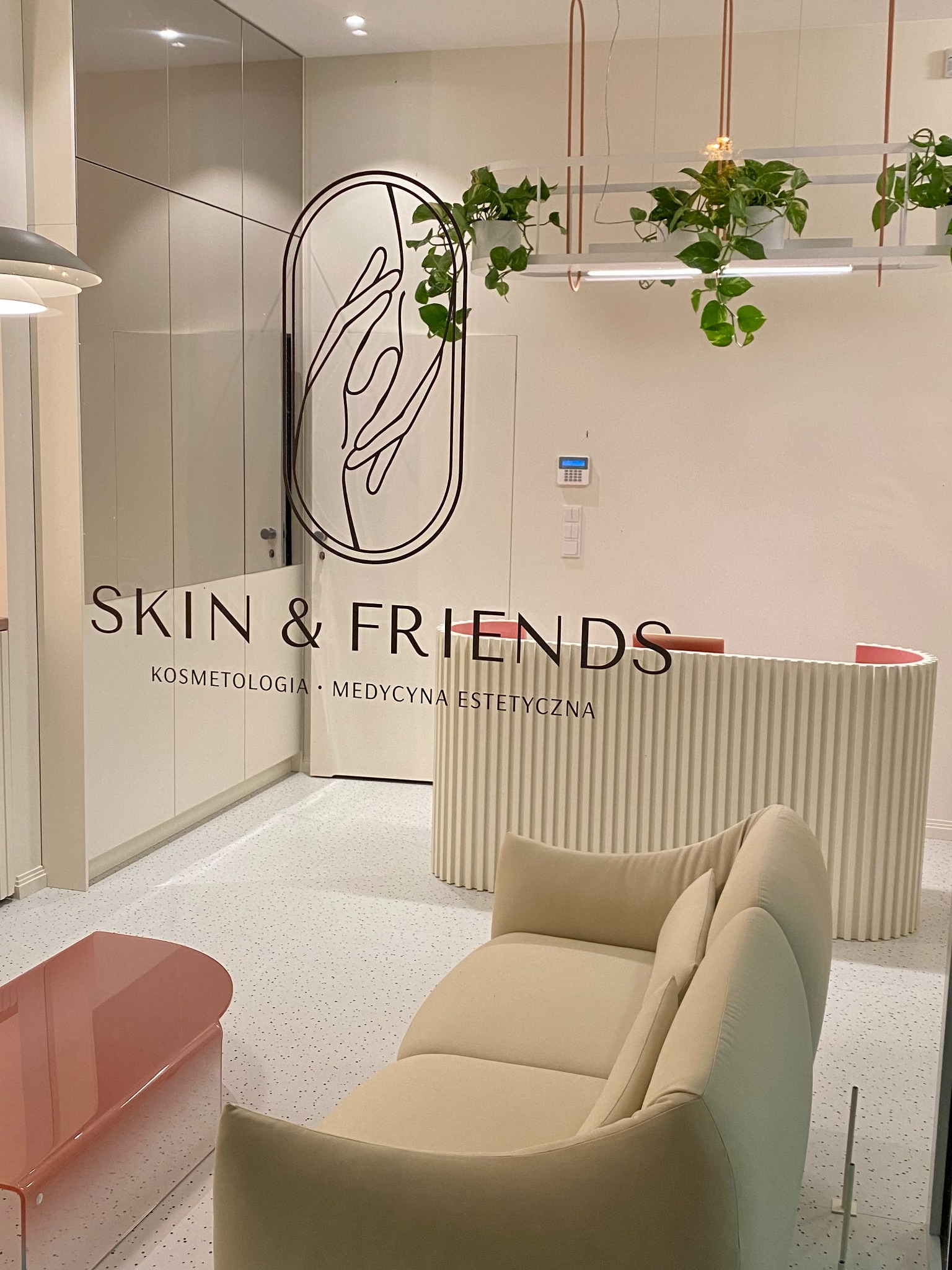 Jasne wnętrze salonu z recepcją, sofą i przeszklonym wejściem z napisem 'SKIN & FRIENDS', dekorowane roślinami.