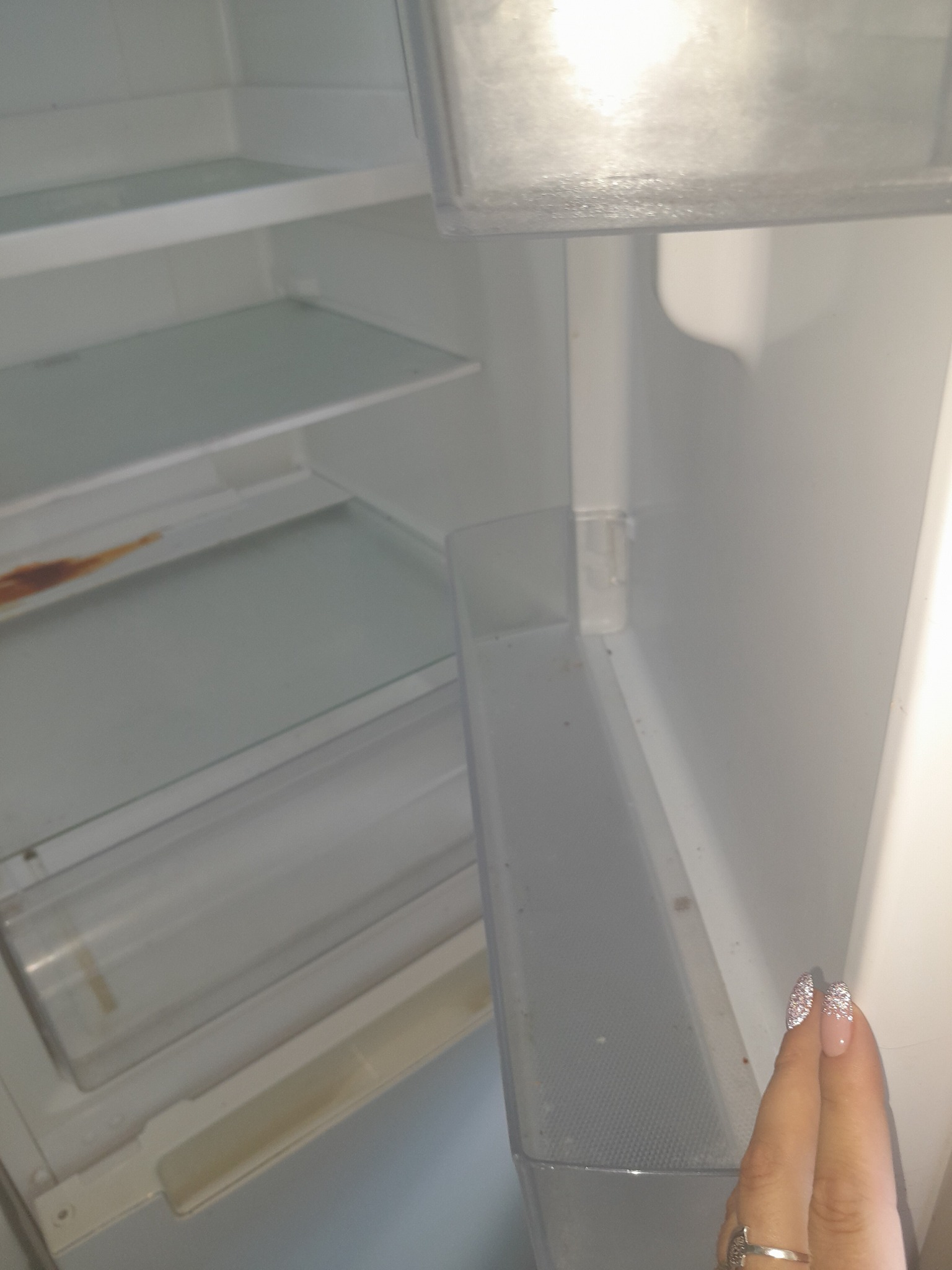 Wnętrze brudnej lodówki z widocznymi plamami i zabrudzeniami na półkach, uchwycone z bliska, z fragmentem dłoni z ozdobnymi paznokciami w prawym dolnym rogu.