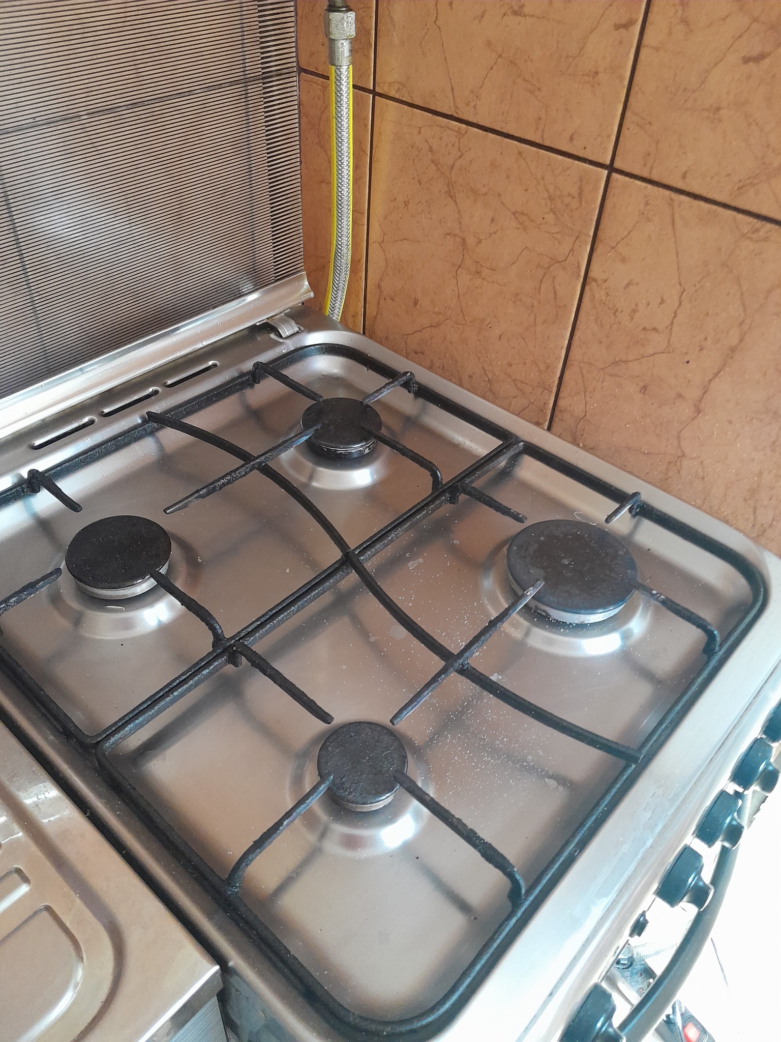 Brudna, czteropalnikowa kuchenka gazowa ze stali nierdzewnej, widoczna z góry pod kątem, z czarnymi rusztami i palnikami, z otwartą klapą osłaniającą ścianę i żółtym przewodem gazowym.