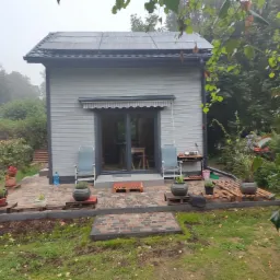 Niewielki, szary domek z panelami słonecznymi na dachu, otoczony zielenią i dekoracyjnym brukowanym tarasem z roślinami w donicach, uchwycony w pochmurny dzień.