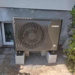 Czarna pompa ciepła marki Kaisai, zamontowana na betonowych bloczkach przy ścianie budynku z małym oknem i pnączem winorośli.