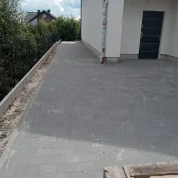 Świeżo ułożona szara kostka brukowa prowadząca do drzwi domu, widoczna paleta drewniana, obrzeża chodnika w trakcie wykończenia, dom w tle.