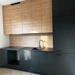 ACTUAL - Nowoczesna kuchnia z czarnymi dolnymi szafkami i drewnopodobnymi górnymi szafkami, czarny blat i armatura, parkiet w jodełkę na podłodze.