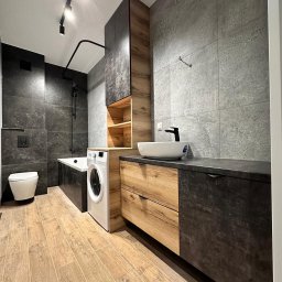 ACTUAL - Nowoczesna łazienka w Toruniu z drewnianymi akcentami: szafka pod umywalkę z szufladami, wysoka szafka z otwartymi półkami, pralka obok wanny, szare płytki na ścianach i drewnopodobna podłoga.