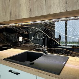 ACTUAL - Czarny zlew z baterią na blacie z dekorem drewna, nad nim szklany panel z abstrakcyjnym złotym wzorem i oświetleniem LED, widoczne białe fronty szafek z czarnymi uchwytami.