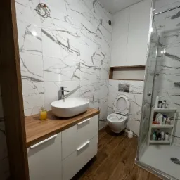 Nowoczesna łazienka z blatem drewnopodobnym pod umywalką nablatową, białymi szafkami, marmurowymi płytkami na ścianach i kabiną prysznicową.