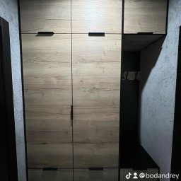 ACTUAL - Szafa wnękowa z drewnopodobnymi frontami i czarnymi uchwytami, z otwartą wnęką na buty i wieszaki, umieszczona w korytarzu o szarych ścianach.