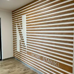 Drewniana dekoracja ścienna z poziomych listew, tworząca logo i napis 'NOWE STAWKI' na białym tle, widok z perspektywy podłogi z płytek imitujących drewno.