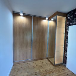 ACTUAL - Wnętrze pokoju z dużą szafą przesuwną z drewnopodobnymi frontami i przeszklonym segmentem z półkami, oświetloną od góry, na drewnianej podłodze.