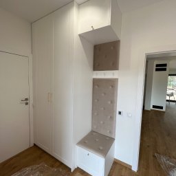 ACTUAL - Biała szafa wnękowa z siedziskiem i panelem tapicerowanym w przedpokoju, widoczne plastikowe zabezpieczenia na podłodze.