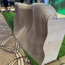 ACTUAL - Falista ścianka działowa z jasnego drewna, wykonana z wielu warstw, ustawiona na sztucznej trawie, z widocznymi śrubami mocującymi, w tle osoby i elementy scenografii.